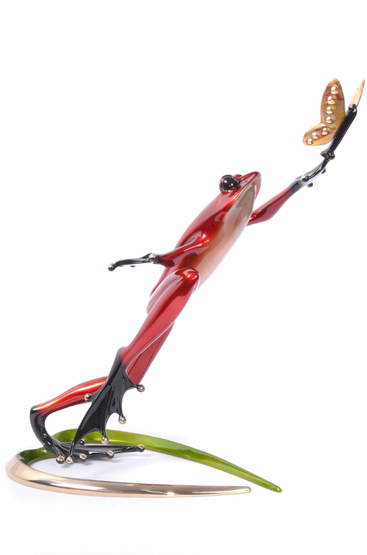 The Be Mine Frogman sculpture features a sleek red frog with black legs and wide eyes, leaping toward a butterfly on its front leg. It is elegantly mounted on a curved green and gold base, capturing vibrant motion.