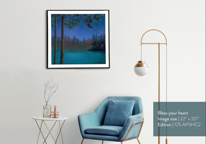 A modern living room showcases a blue armchair, a small round table with a vase of delicate branches, and a gold floor lamp. On the light-colored wall hangs Bless Your Heart, a framed landscape painting reminiscent of Mackenzie Thorpes signature pastel on paper style.