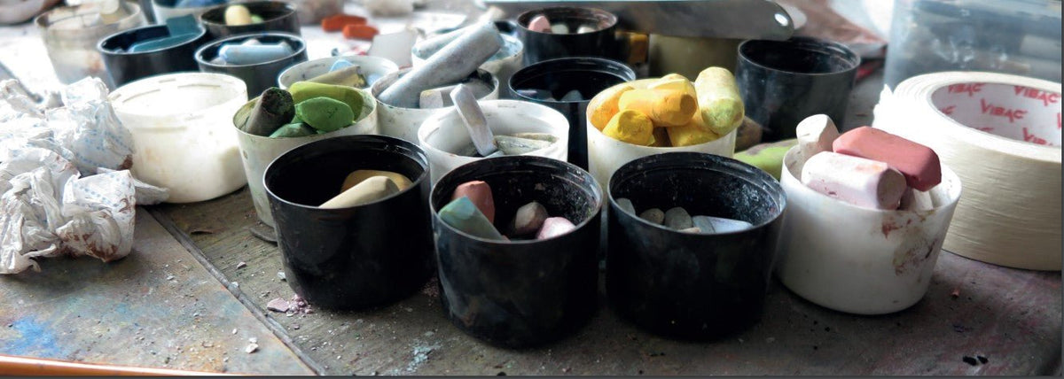 Containers of colorful chalk and blending tools are arranged on a paint-splattered table, alongside crumpled tissues and tape, revealing an artists process reminiscent of Mackenzie Thorpes Bless Your Heart style with pastel on paper.