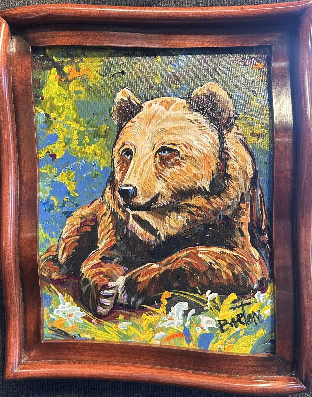 Bleu by Steve Barton is a colorful wildlife painting featuring a brown bear resting on grass among flowers, set against green and yellow foliage. The vibrant artwork, signed BARTON, comes displayed in a wooden frame.