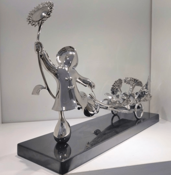 The Bringer of Sunshine-Stainless by Mackenzie Thorpe is a limited edition silver sculpture featuring a stylized figure with a sunflower and a cart full of sunflowers, displayed on a sleek black rectangular base.