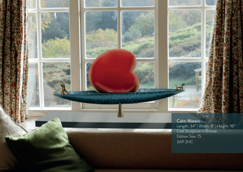 Mackenzie Thorpes Calm Waters, a kidney-shaped red sculpture on a blue, boat-like base with two small figures, is showcased at Marcus Ashley Gallery in front of a scenic window highlighting its details.