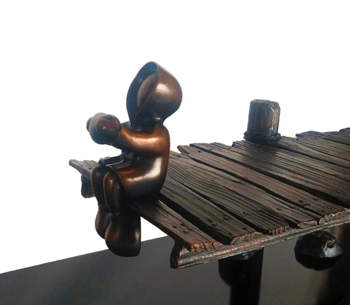 A small bronze figure by Mackenzie Thorpe, titled Dock of the Bay, sits hooded on a rustic wooden dock, holding something in its hands. The limited edition sculpture stands out against the plain, light background.