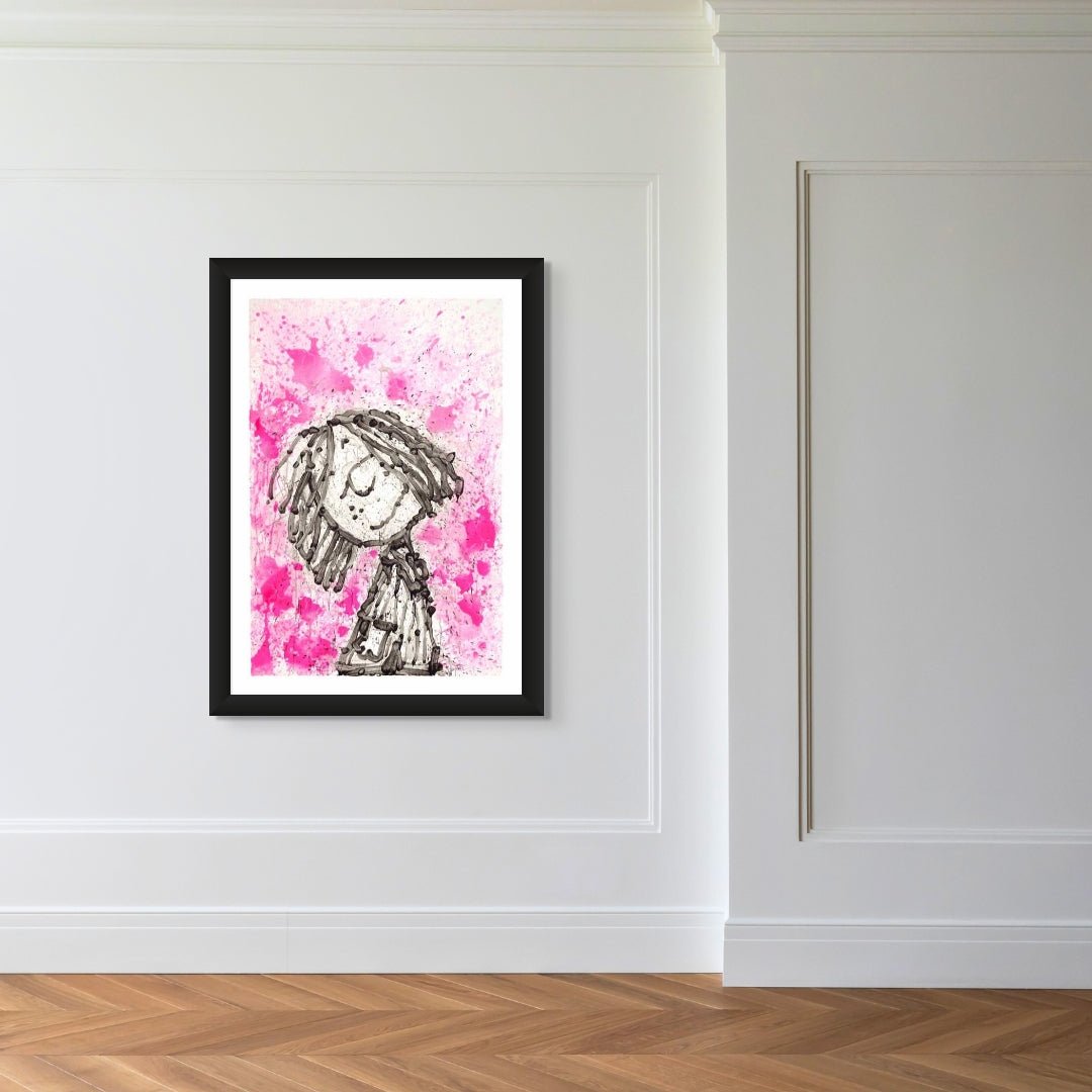 The Home Girl Dreams framed painting, featuring a whimsical child with textured hair in a style reminiscent of Tom Everhart, hangs on a minimalist white wall above a hardwood floor against a vibrant pink and white background.