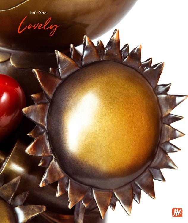 A close-up of sun-shaped metallic sculptures in golden and bronze hues, surrounded by red cherries. Isnt She Lovely is written in the top left corner, capturing emotional resonance. A red logo at the bottom right complements this modern art piece.