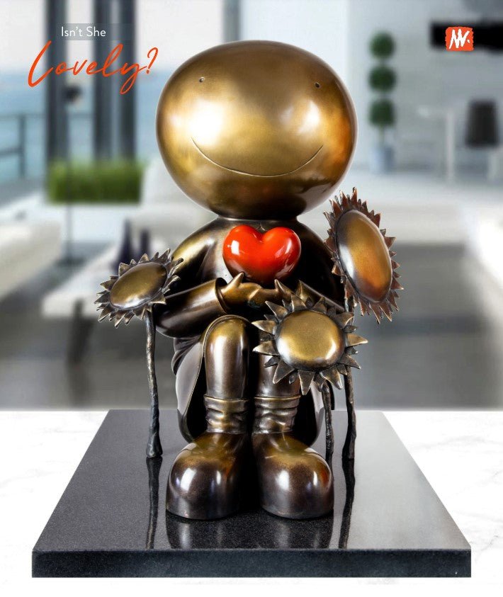 The metallic sculpture Isnt She Lovely by Mackenzie Thorpe features a cross-legged, round-headed figure holding a red heart and smiling amidst sunflower-like elements, set against a blurred modern interior that highlights its emotional depth and contemporary essence.