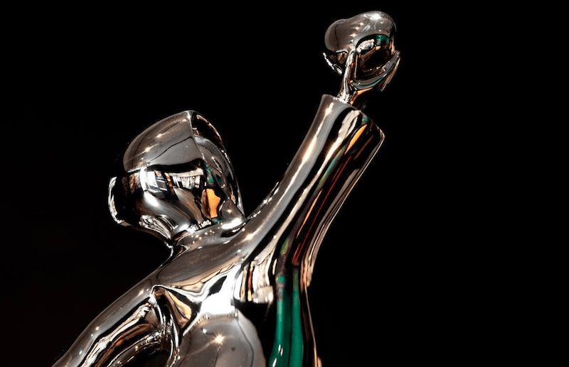 A close-up of Love Picker - Stainless reveals a shiny, abstract silver sculpture against a black backdrop. This limited edition piece boasts fluid metallic forms with dynamic curves and an open top structure, subtly reflecting light and colors akin to Mackenzie Thorpes artistic style.