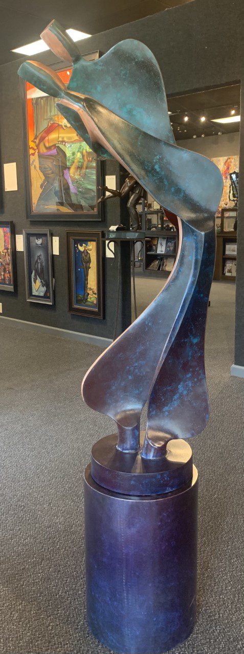 The Tango - Copper sculpture, with its sleek and abstract interlocking curves, rises from an 18 base. Surrounded by framed paintings in the art gallery, it captivates viewers, while the dim lighting enhances the contemporary ambiance.