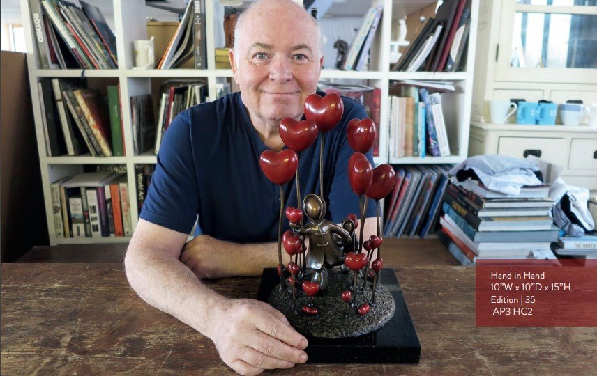 A person wearing a blue shirt smiles while sitting at a table featuring the Hand in Hand sculpture surrounded by red heart-shaped balloons. Books and diverse objects adorn the background shelves.