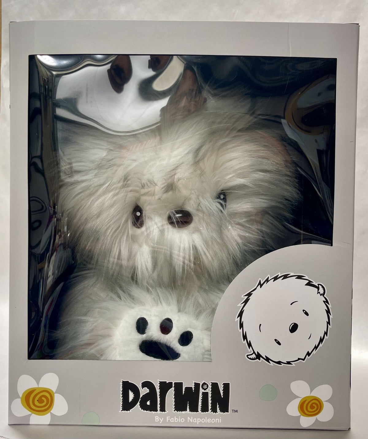 The Darwin Plush, by Fabio Napoleoni, is a fluffy white toy resembling a dog or bear with button eyes and a small nose. It comes nestled inside a collectors box adorned with flowers and character illustrations, making it ideal for fans and collectors.