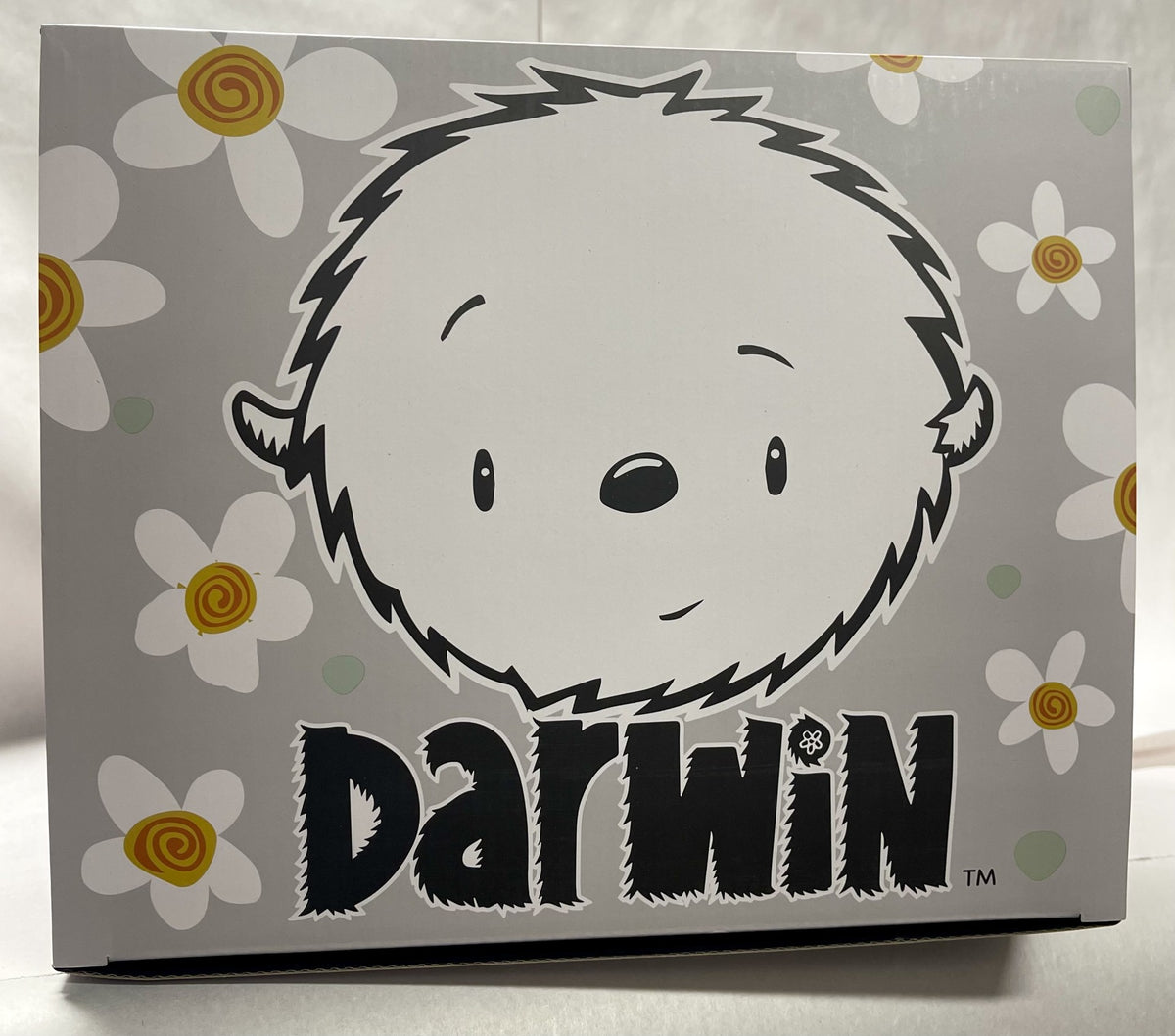 A collectors box with the cartoon face of a fluffy, round-faced character is surrounded by white and orange flowers. Darwin Plush is printed below in bold, stylized letters, with a light gray background enhancing the playful design reminiscent of Fabio Napoleonis artistry.