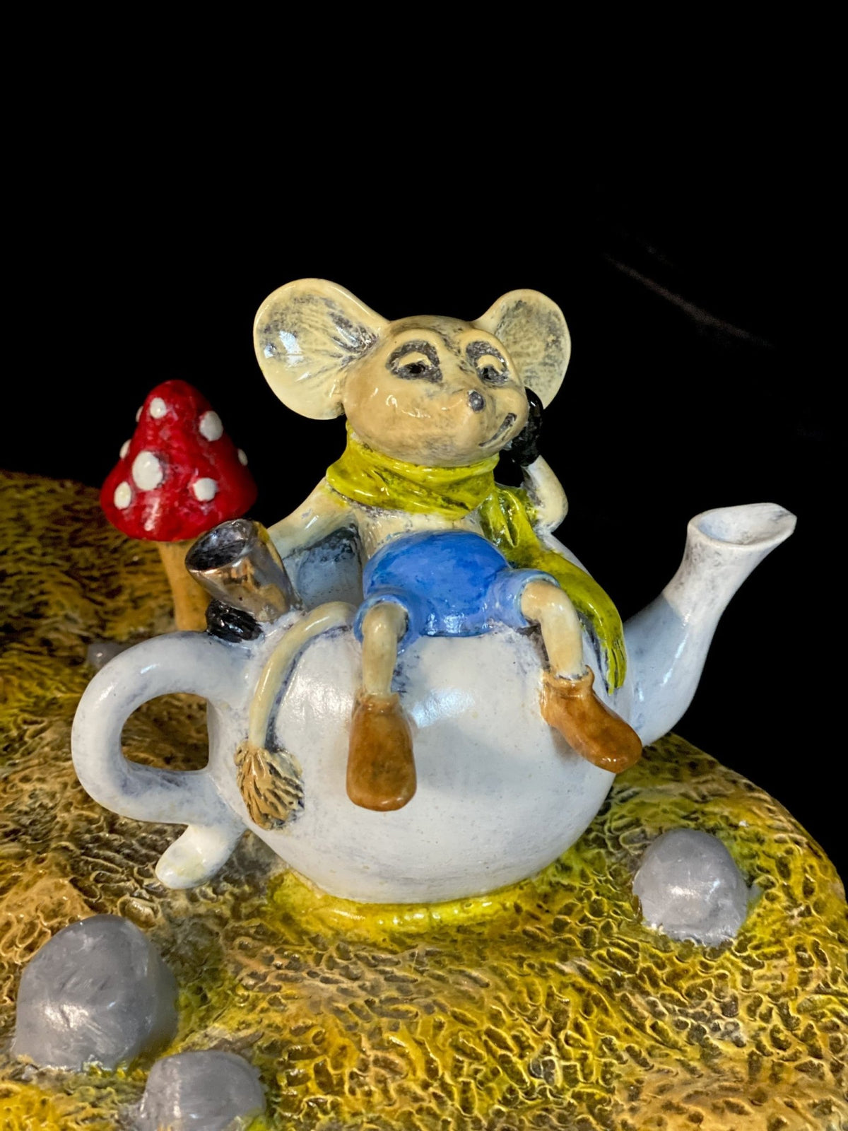 The White Rabbit, a whimsical ceramic figurine inspired by Alice’s adventures, features a green scarf and blue shorts on a white teapot, with a red-and-white mushroom beside it on a textured yellow surface with gray stones.