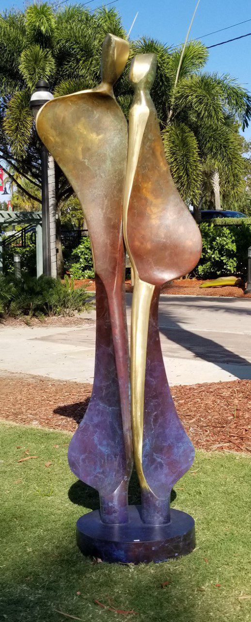 The Quiet Encounter - Brass is a tall, abstract sculpture of two slender figures standing side by side, crafted from smooth metal. Their colors transition seamlessly from bronze to purple, set gracefully in a grassy area framed by palm trees.