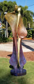The Quiet Encounter - Brass is a tall, abstract sculpture of two slender figures standing side by side, crafted from smooth metal. Their colors transition seamlessly from bronze to purple, set gracefully in a grassy area framed by palm trees.