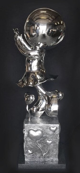 The shiny silver sculpture My Butterfly-Stainless by Mackenzie Thorpe features a child figure with a large round head on a platform, holding one hand up. Heart-shaped designs decorate its base, and it has a polished reflective surface. This sculpture is a limited edition piece.