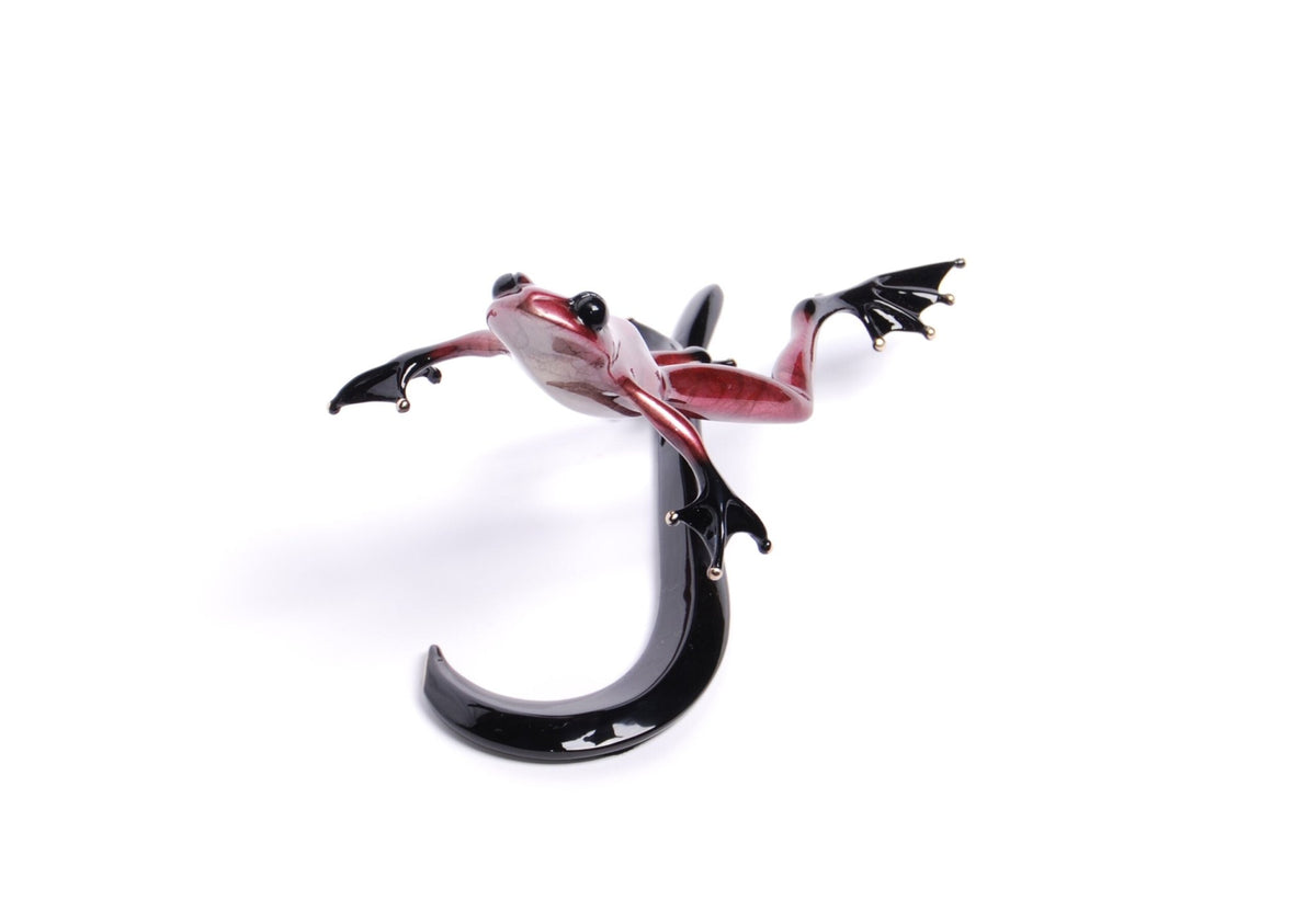 Valentino is a glass sculpture of a red frog with a black body, artfully posed with outstretched legs and elegantly curved limbs and tail, leaping against a white background, capturing the essence of motion.