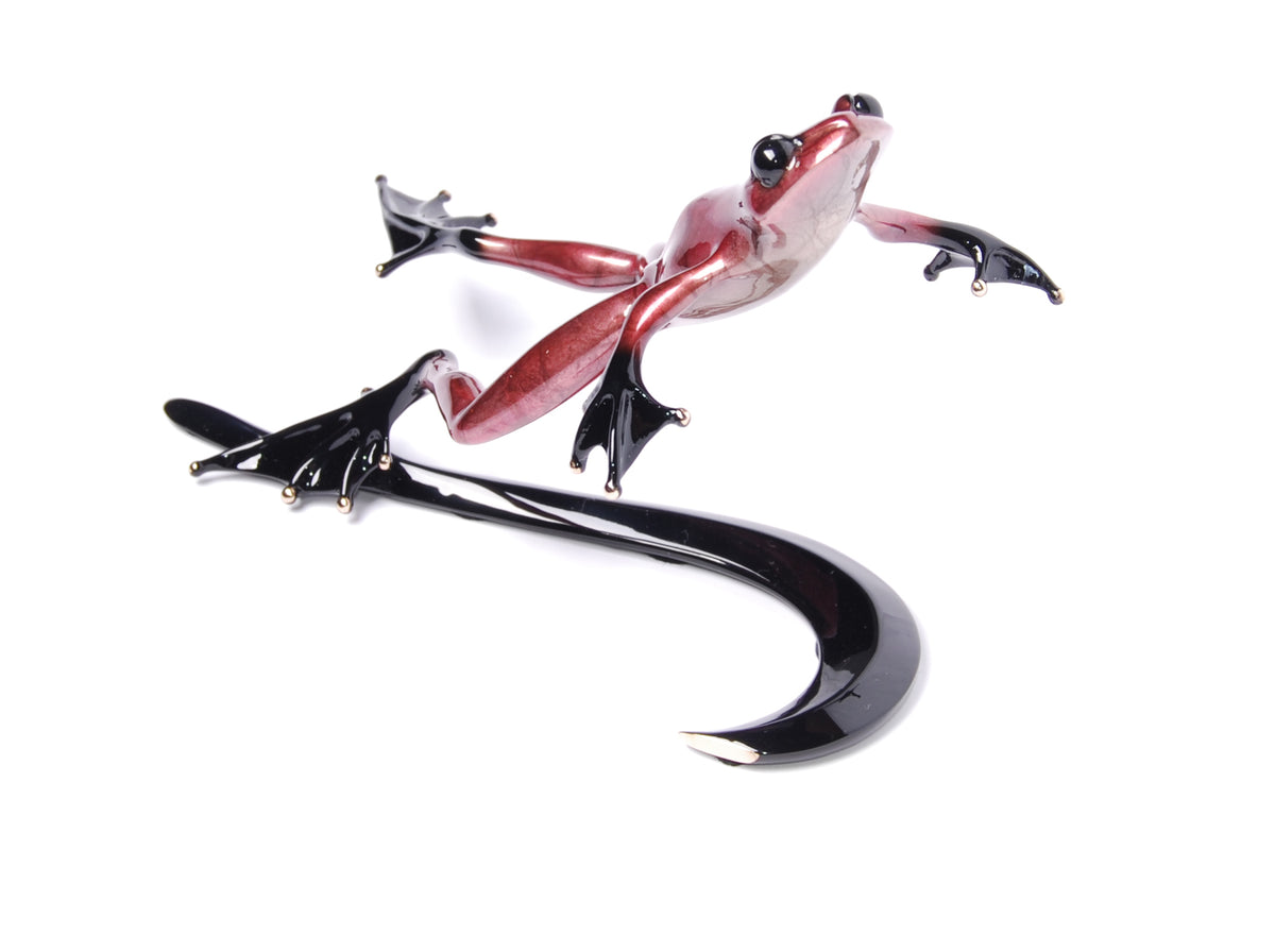 Part of the Valentino by Frogman Bronze collection, this shiny, stylized leaping frog sculpture features outstretched limbs and a striking red frog patina. It perches on a long, curved black structure resembling a fish hook and is set against a white background.